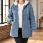 A woman wears a blue hooded jacket with plush texture, a cream lined hood and front zipper.