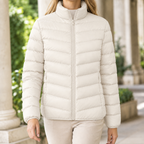 A woman wears an off white quilted puffer jacket with chevron quilting, a stand collar and front zip.