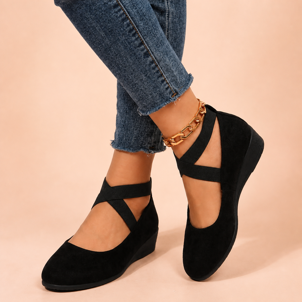 Black suede flats on feet with crossed legs in blue jeans and gold anklet